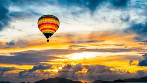 Bunter Heißluftballon schwebt über einem See bei Sonnenuntergang mit dramatischem Wolkenhimmel.