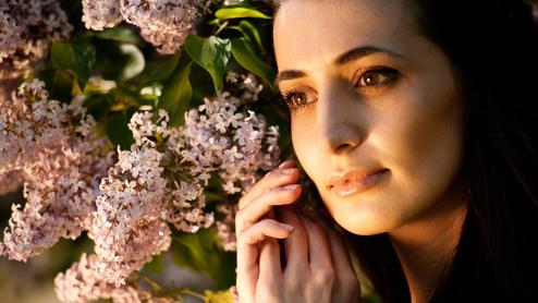 Nahaufnahme einer Frau mit langen, dunklen Haaren, die sanft von lila Fliederblüten umgeben ist.